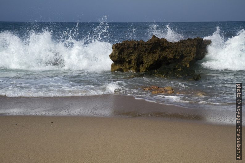 Vague qui éclate derrière un rocher sur la plage. Photo © André M. Winter